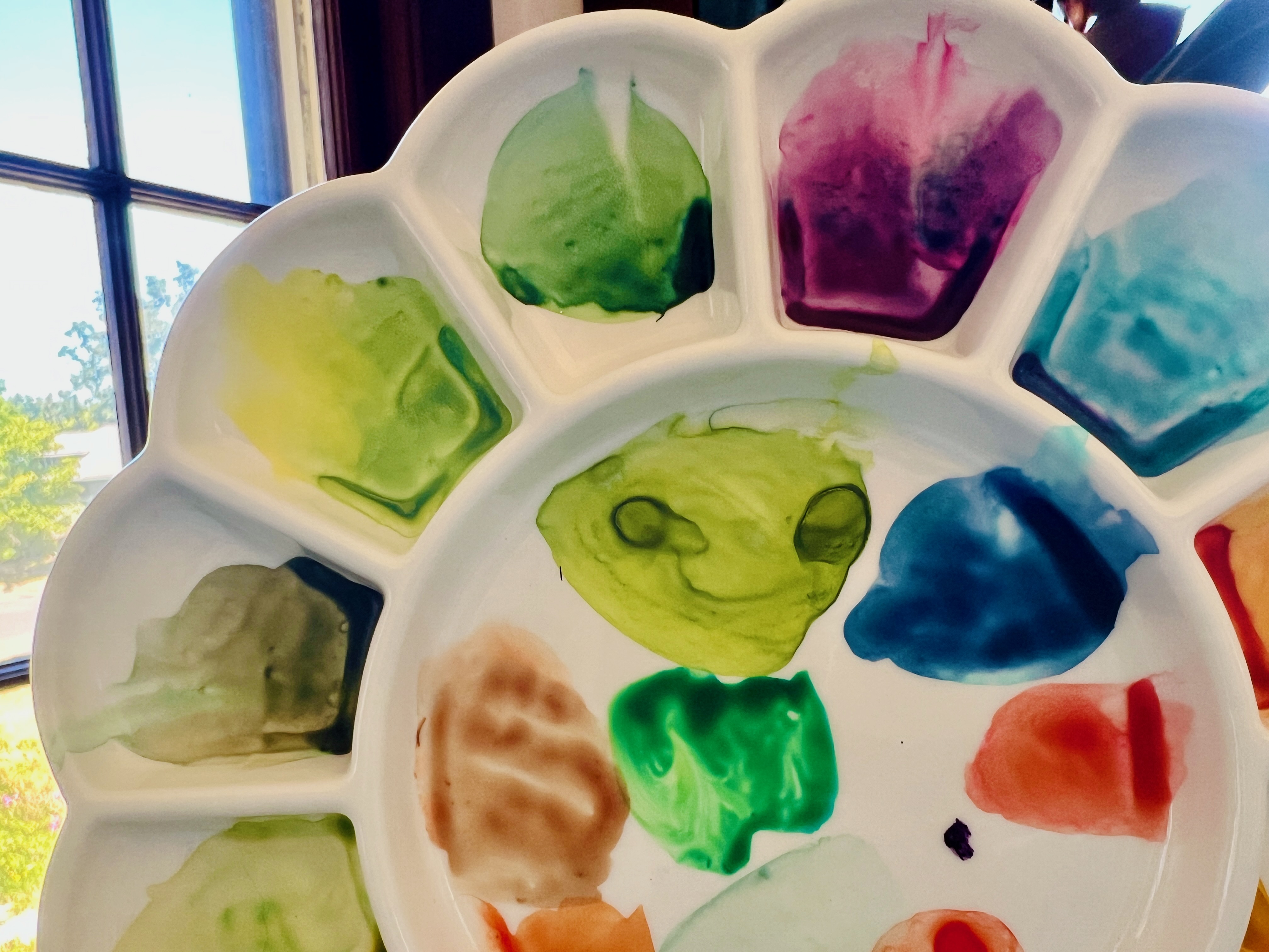 A watercolor paint palette, white ceramic and covered in dabs of paint, and one of them has dried to look like the face of a cute smiling green alien.