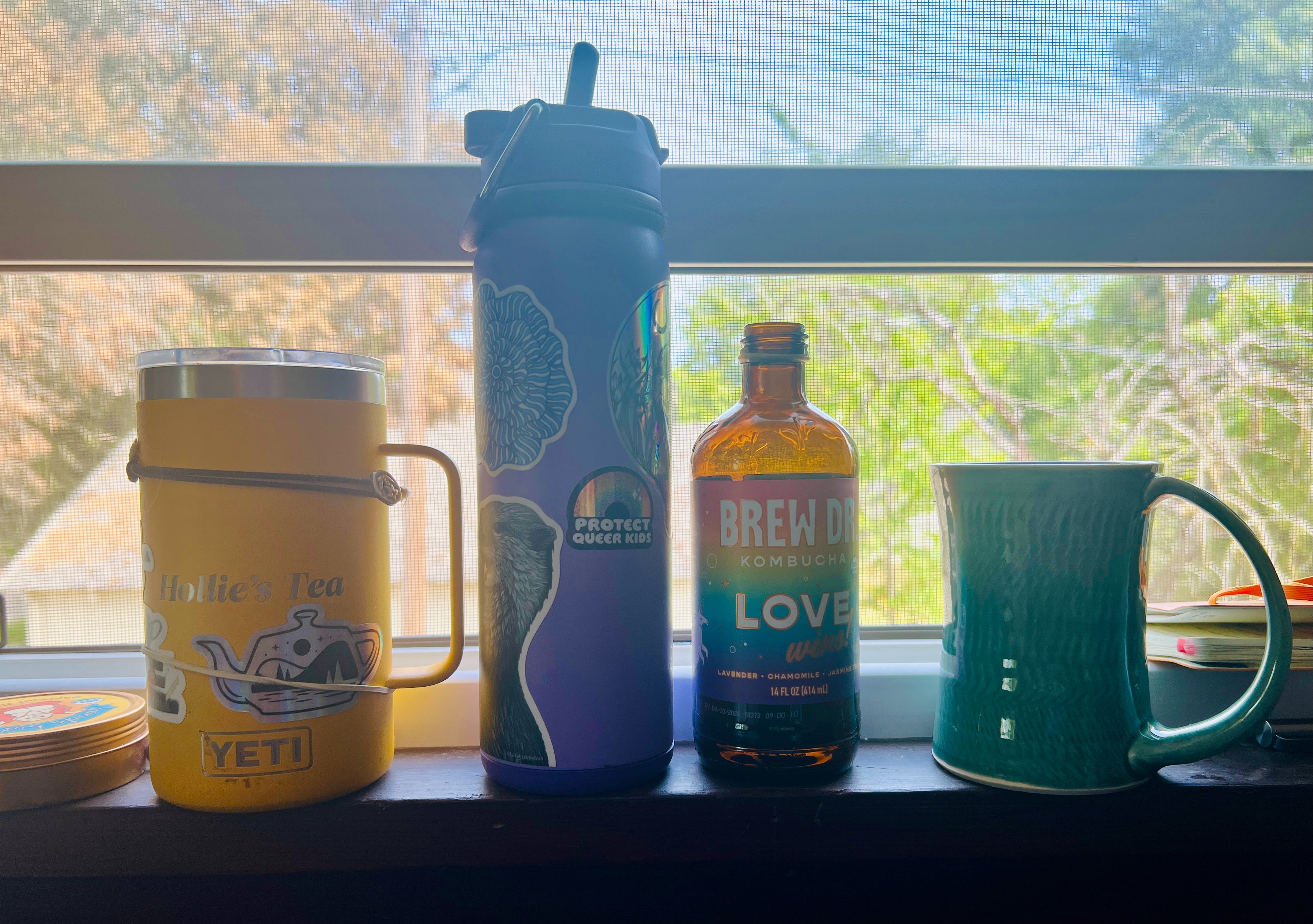 A row of drinks on a windowsill, from L-R: a mug of tea, a purple water bottle, a bottle of kombucha, and another mug of tea.