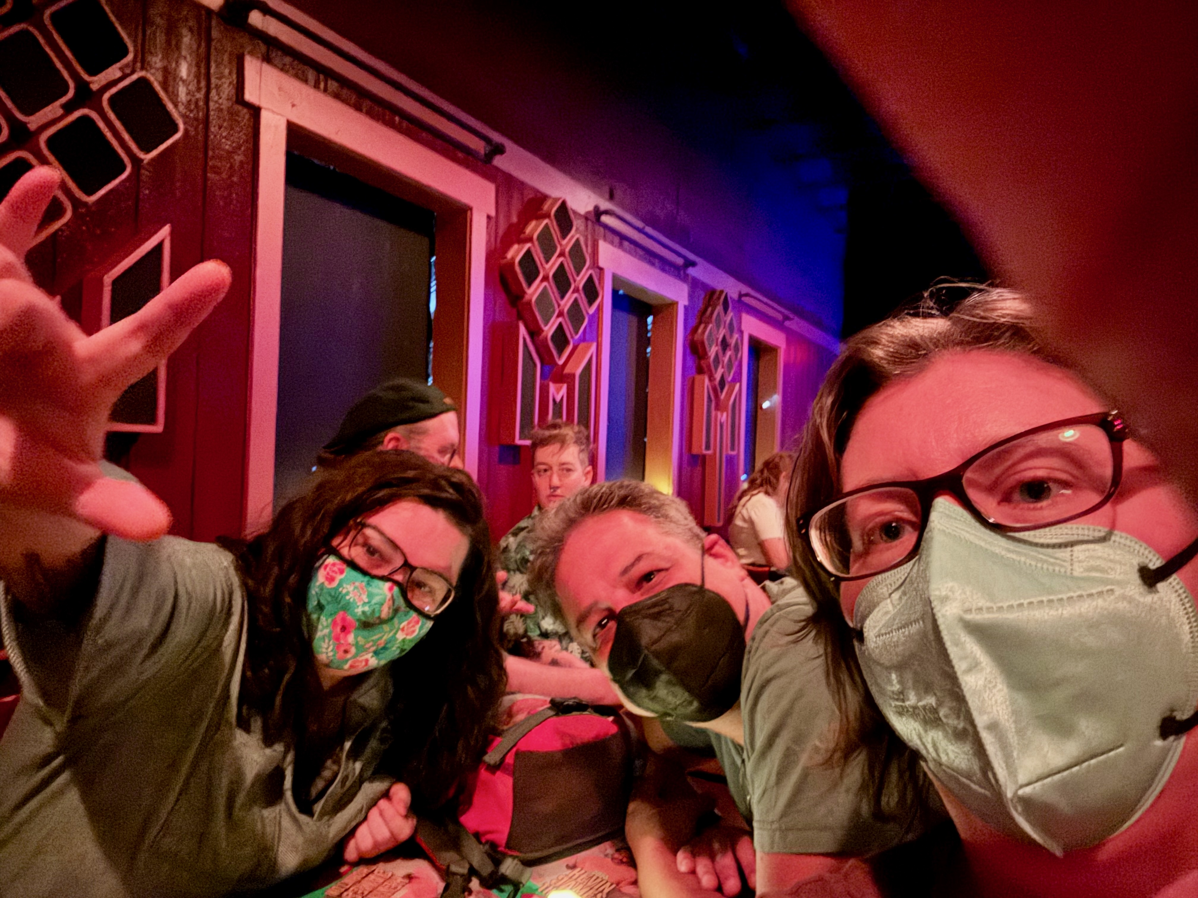 Three people leaning over a table, getting a selfie, in a dark comedy club. They're wearing masks and smiling, though you can't tell.