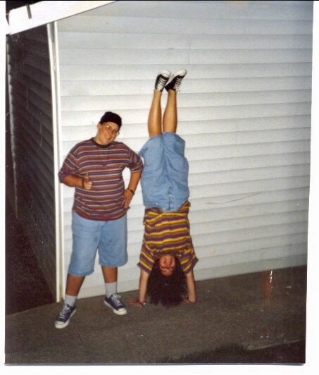 A 15 yo boy standing outside against the wall of a parking structure in an apartment complex. He's wearing denim shorts and a striped shirt. Next to him, a 16yo girl wearing his same outfit, does a handstand against the wall.