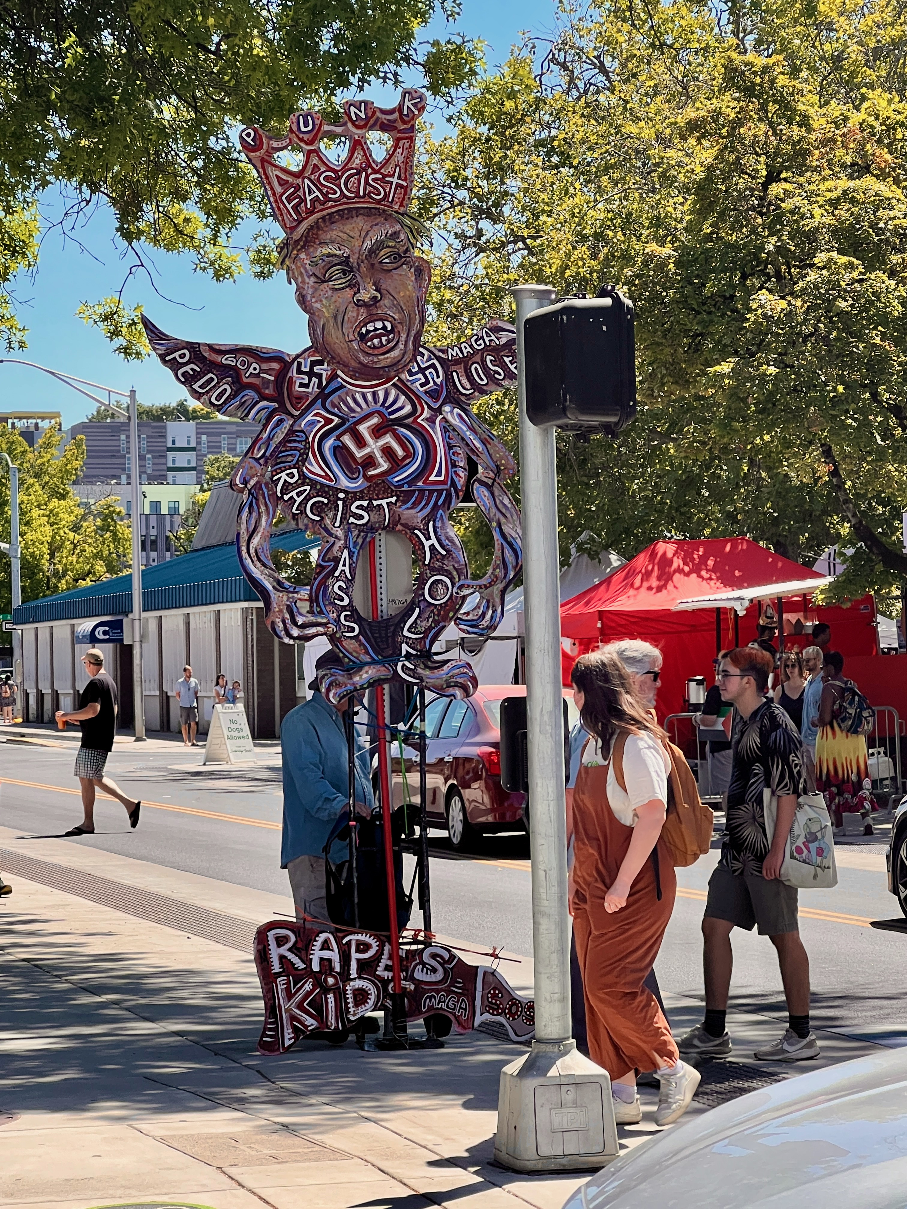 Sunny corner of a busy city block, a large effigy of Trump is held up, with writing all over him detailing his many faults